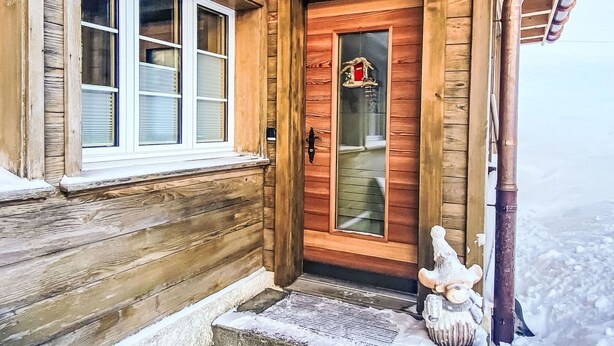 L'ingresso di una baita di legno con una porta in vetro, un piccolo portico e terreno innevato.