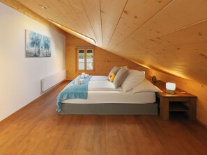 A bedroom with a wooden ceiling, a bed with pillows and a blanket, a nightstand with a lamp, and a window with a view.