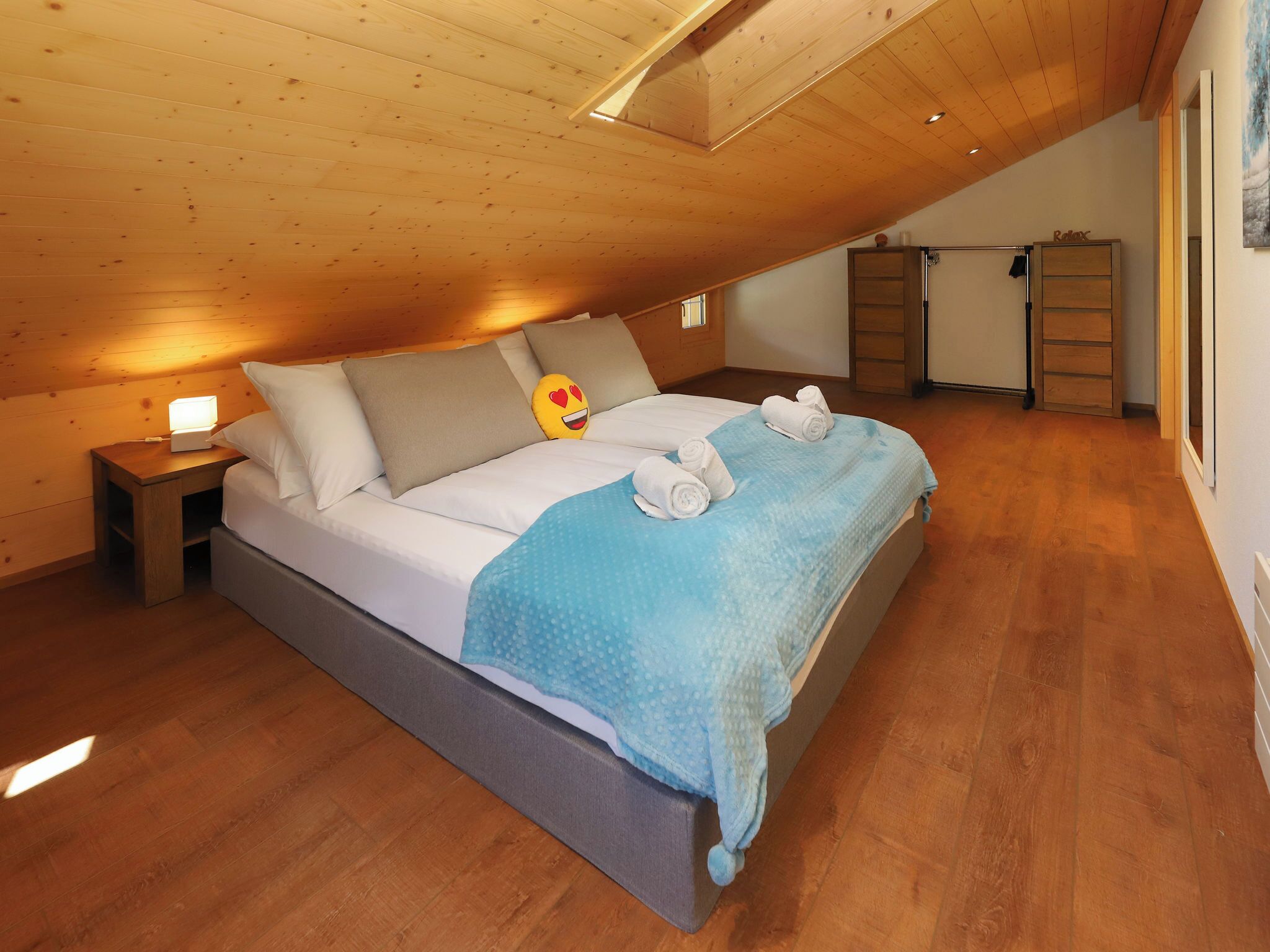 A bedroom with a sloped ceiling, a bed with pillows and a heart-shaped pillow, a nightstand with a lamp, and a blue towel on the bed.