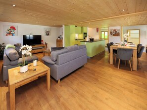 Una sala moderna con un sofá gris, una mesa de centro de madera y un televisor. Se ve una cocina con gabinetes verdes y un comedor con una mesa y sillas de madera.