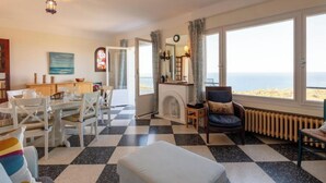 Una habitación con piso de cuadros, una chimenea, un área de comedor con mesa y sillas, un ventanal con vista al mar y un rincón acogedor con un sillón y reposapiés.
