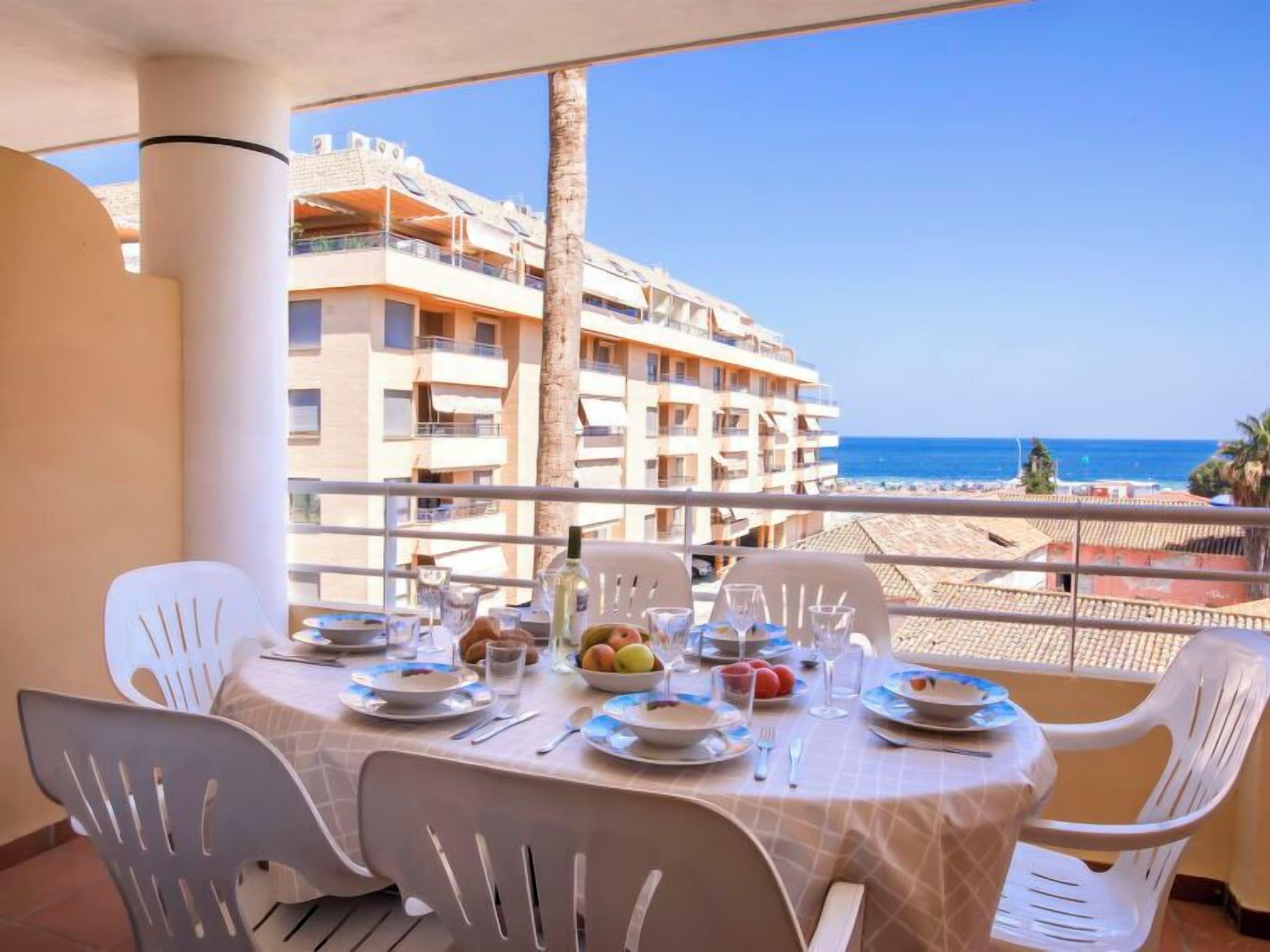 Ein Balkon mit Tisch und Stühlen, gedeckt mit Tellern, Gläsern und Obst, mit Blick auf einen Strand und ein mehrgeschossiges Gebäude.