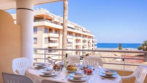 Une table dressée sur un balcon, avec des assiettes, des verres et des fruits, d’où l’on peut admirer une plage et un immeuble à plusieurs étages.