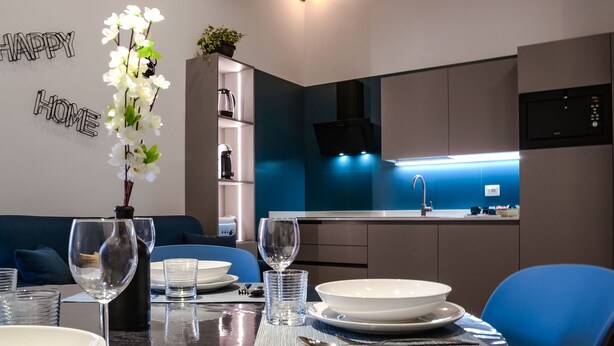 A modern kitchen with a blue and white color scheme, a dining area with blue chairs, and a decorative white orchid plant. - Lele Rooms Termini (Rome)