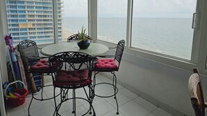 Ein Balkon mit rundem Tisch und Stühlen, einem Blumentopf und Blick auf das Meer und ein Hochhaus.