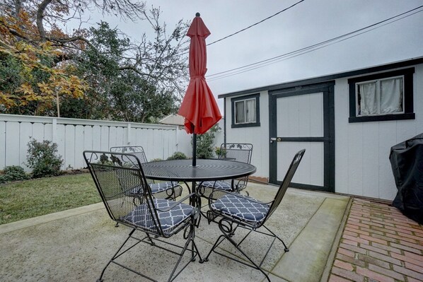 A patio with a round metal table and four chairs, a red umbrella, and a white fence.