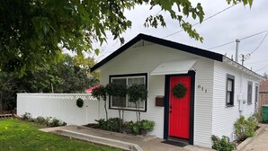 Una pequeña casa de un solo piso con puerta roja, revestimiento blanco y buzón.