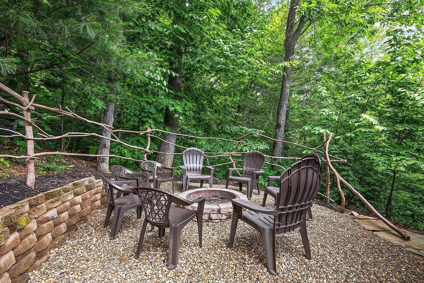 An outdoor seating area with Adirondack chairs and a fire pit surrounded by a gravel path, set in a wooded area.