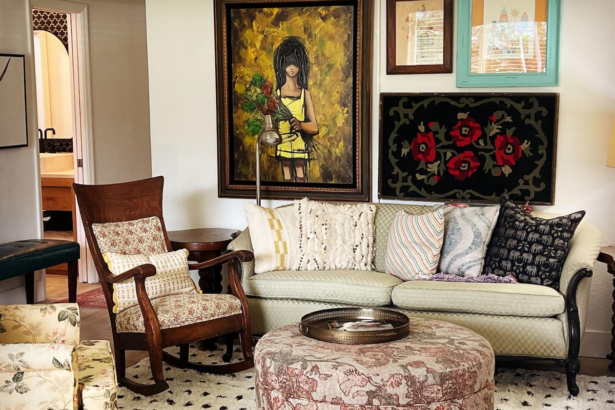 Ein Wohnzimmer mit einer Couch, einem Hocker und einem Schaukelstuhl. An den Wänden hängen Bilder und auf einem Beistelltisch steht eine Lampe.