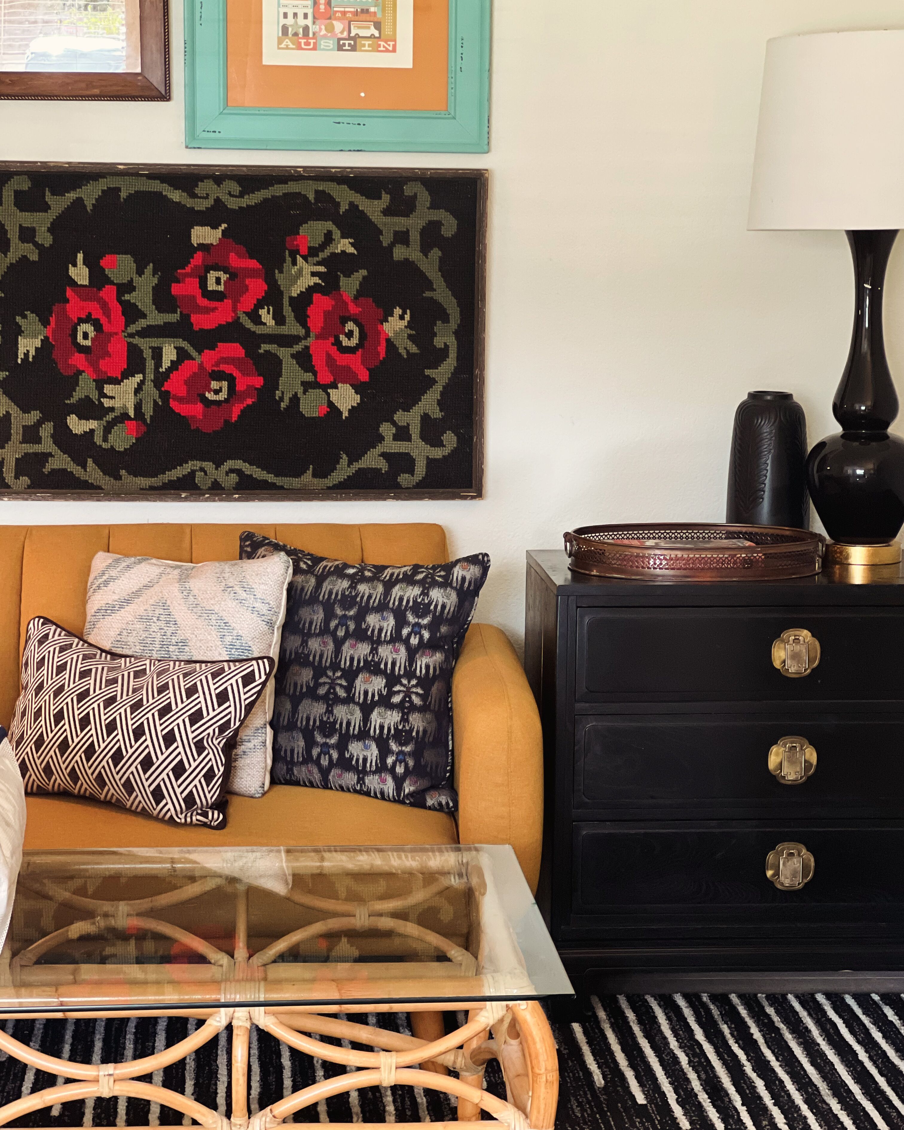 A living room with a sofa, patterned cushions, a glass coffee table, a black dresser, and a framed artwork on the wall.