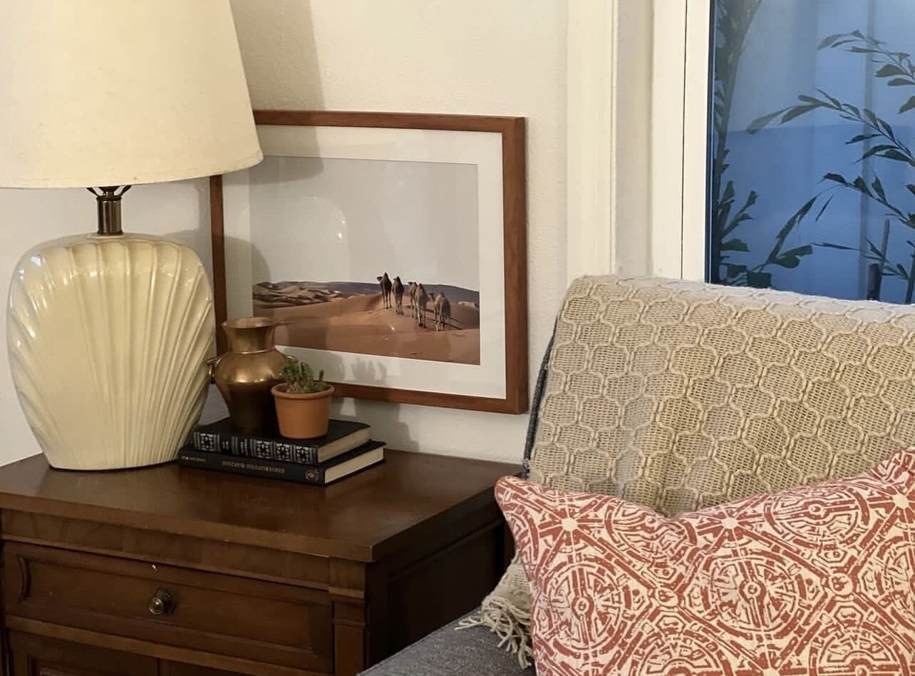 A room with a wooden nightstand, a lamp, a framed picture, a patterned sofa, and a window with a bamboo view.