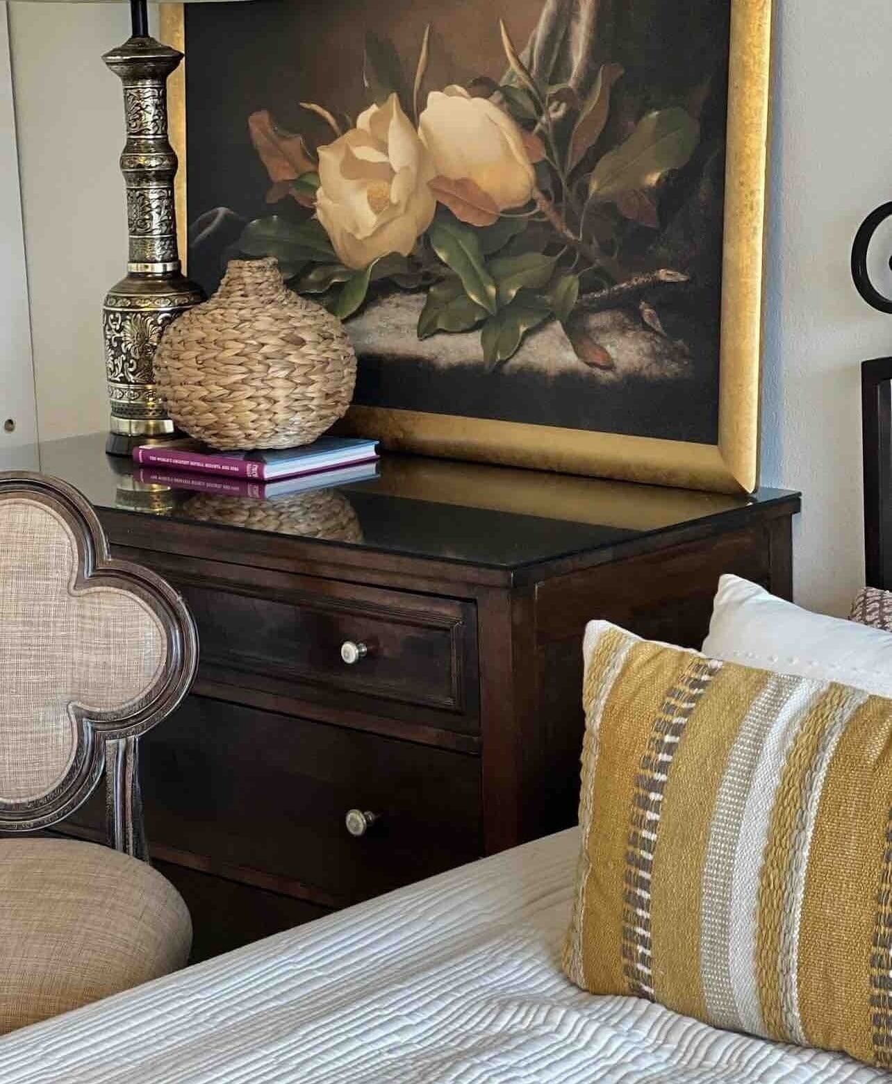 A bedroom with a bed, a nightstand with a lamp, a woven basket, and a framed painting.