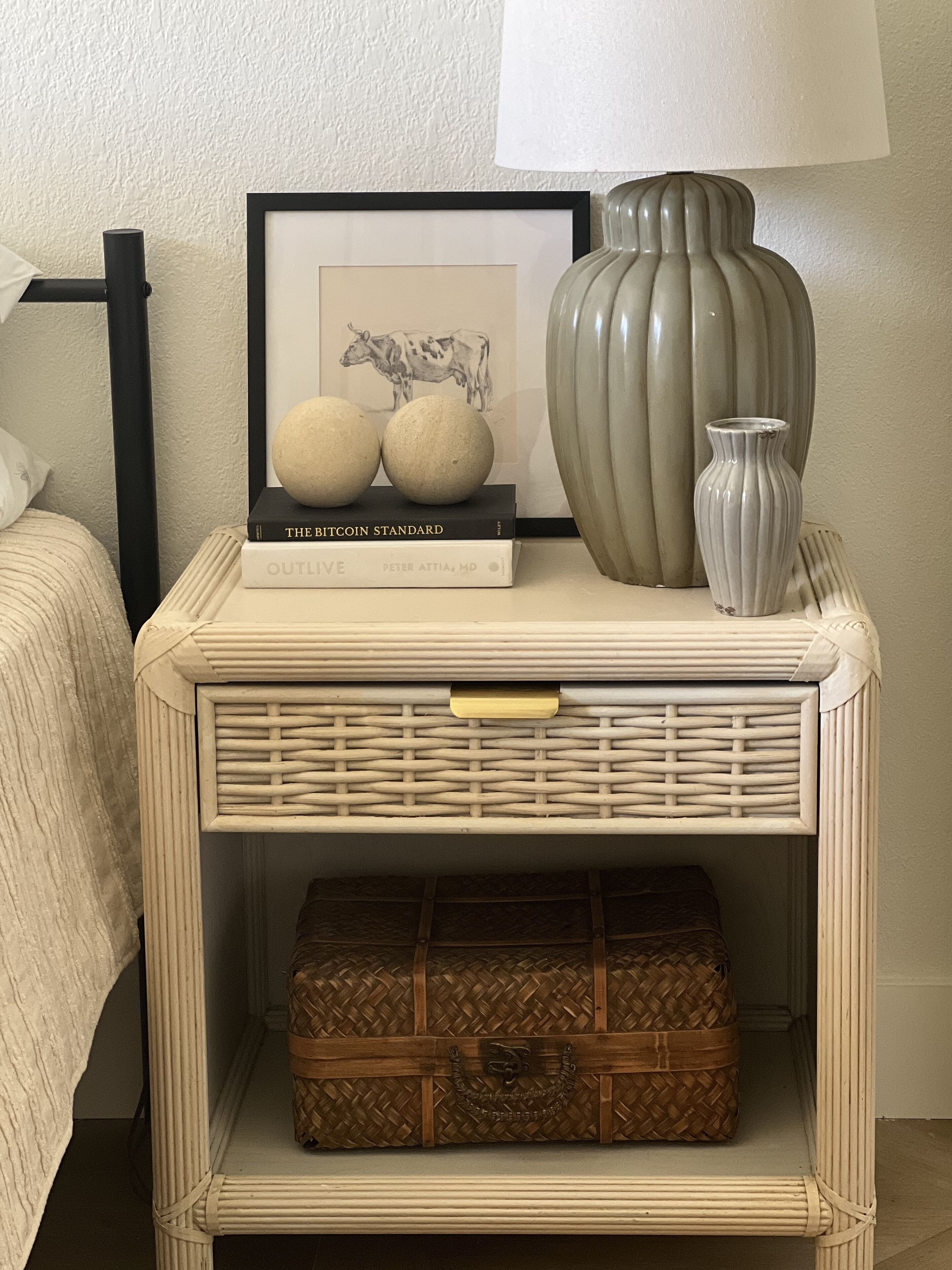 A bedside table with a framed picture, two spherical objects, a vase, and a woven basket.