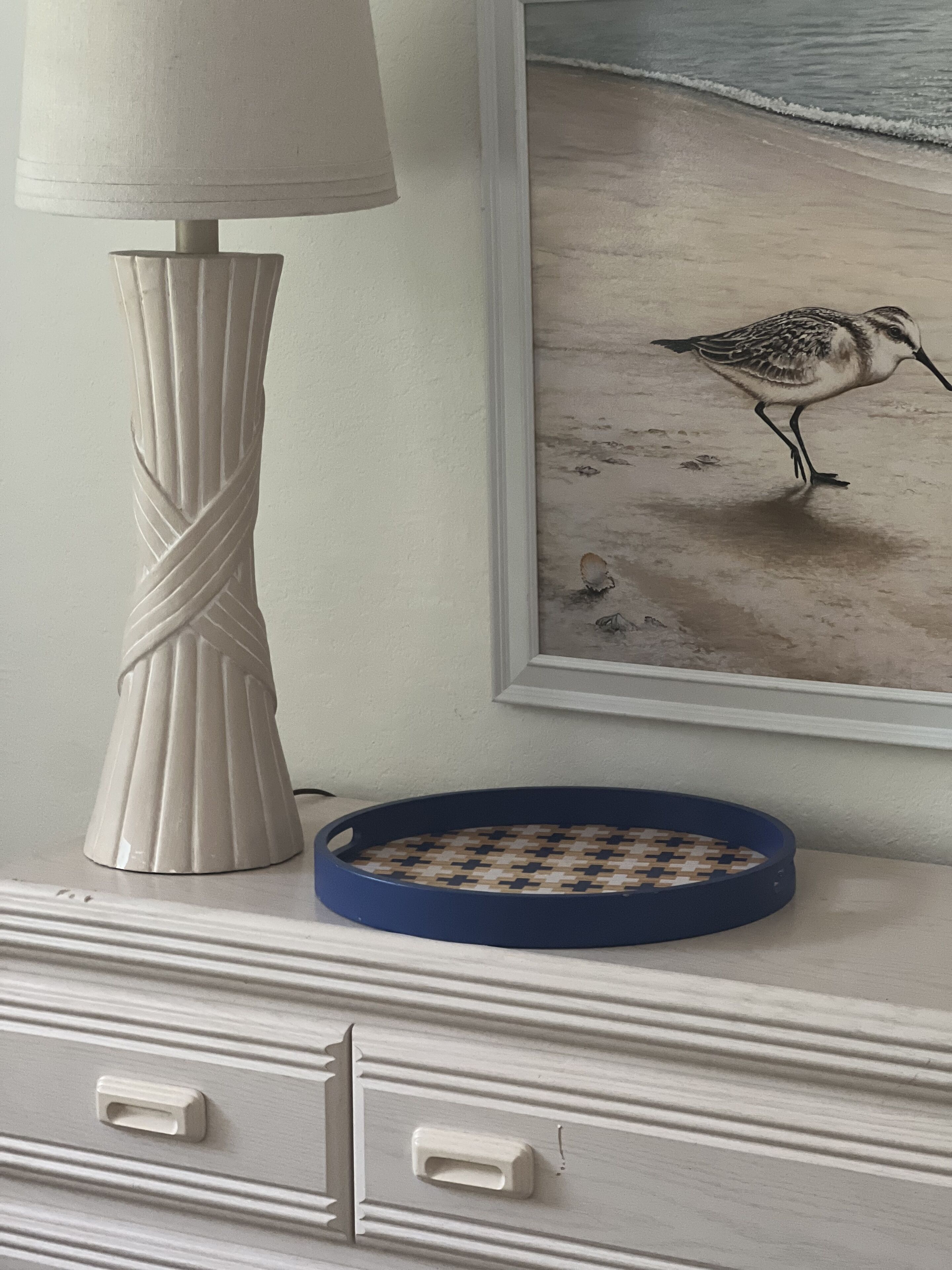 A white vase, a framed bird painting, and a blue tray on a dresser.