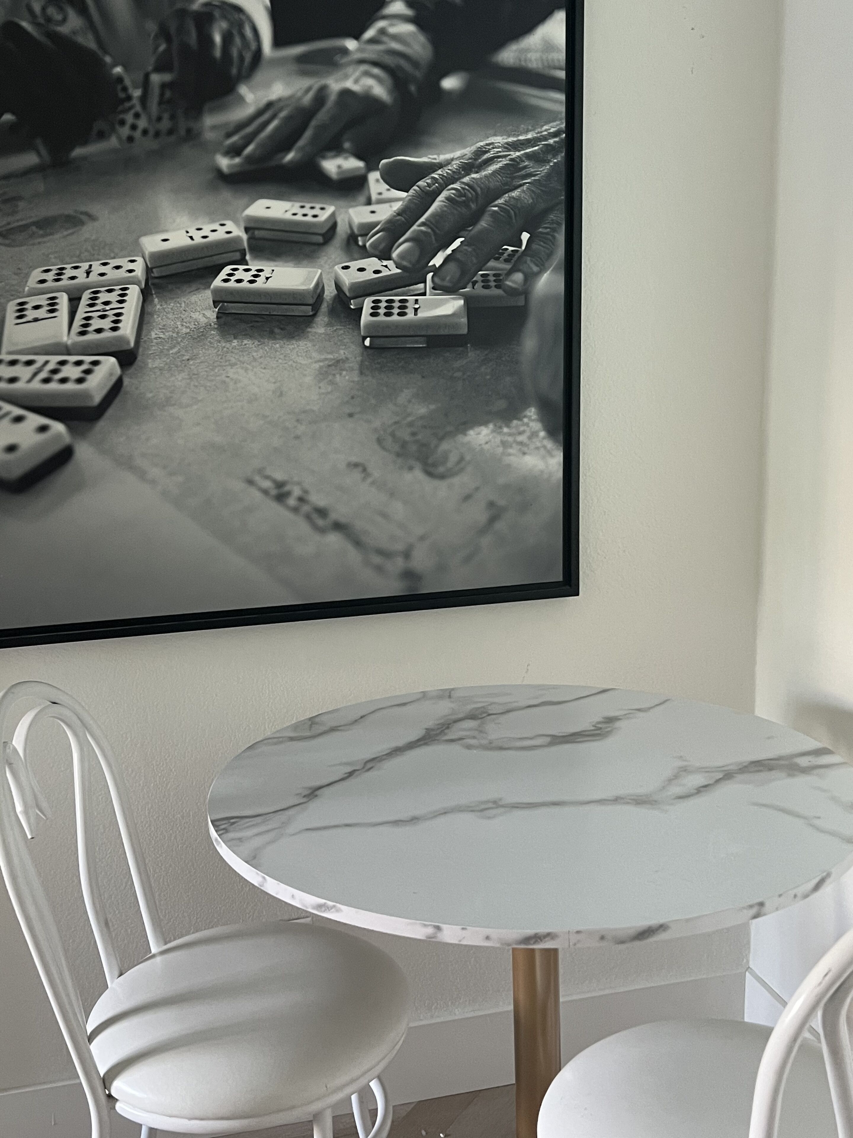 A table with a marble top and two white chairs, positioned in front of a wall-mounted picture of a person playing with dominoes.