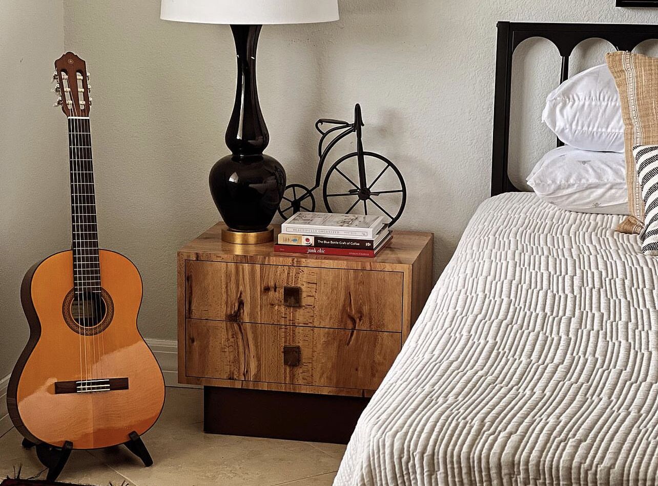 Un dormitorio con mesita de noche de madera, una guitarra, una lámpara, una bicicleta y una cama con ropa de cama a rayas.