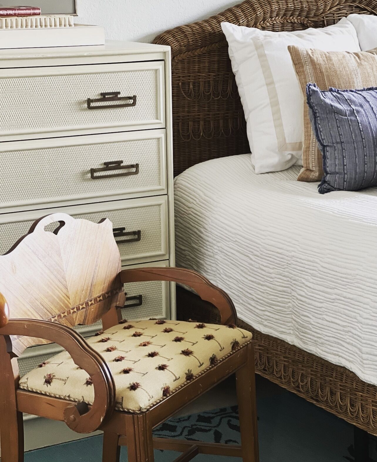Un dormitorio con una cama, una silla de madera con cojín, un aparador blanco con cajones y un cabecero de mimbre.