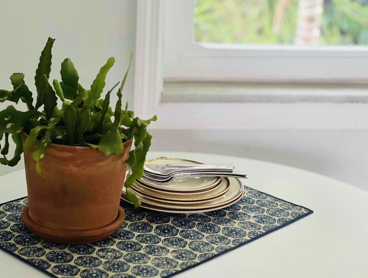 Una planta en maceta sobre un mantel estampado, con platos apilados y un tenedor.