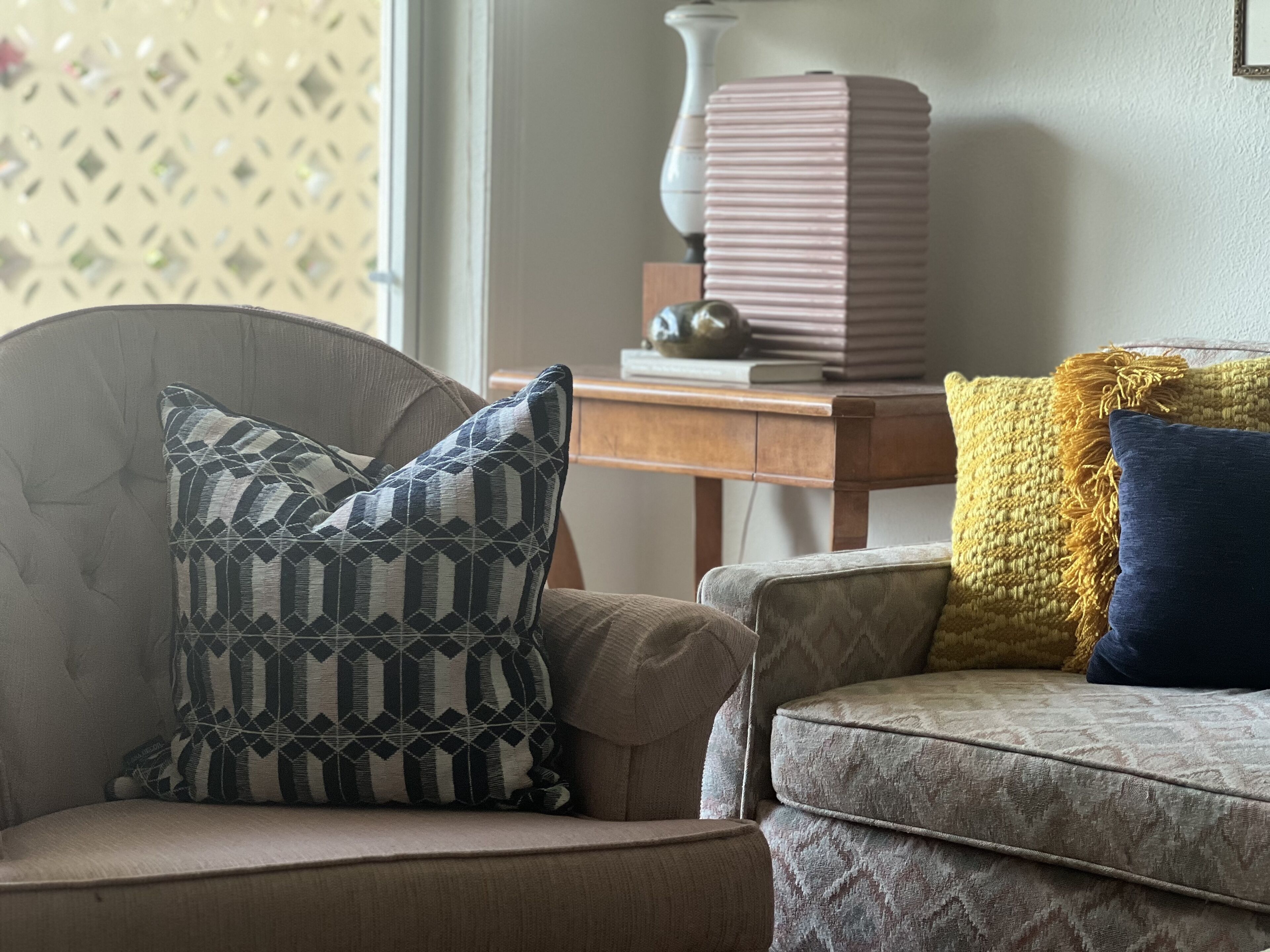 Uma sala de estar com uma poltrona estampada, um sofá com almofadas decorativas e uma mesa lateral de madeira com uma luminária e livros.