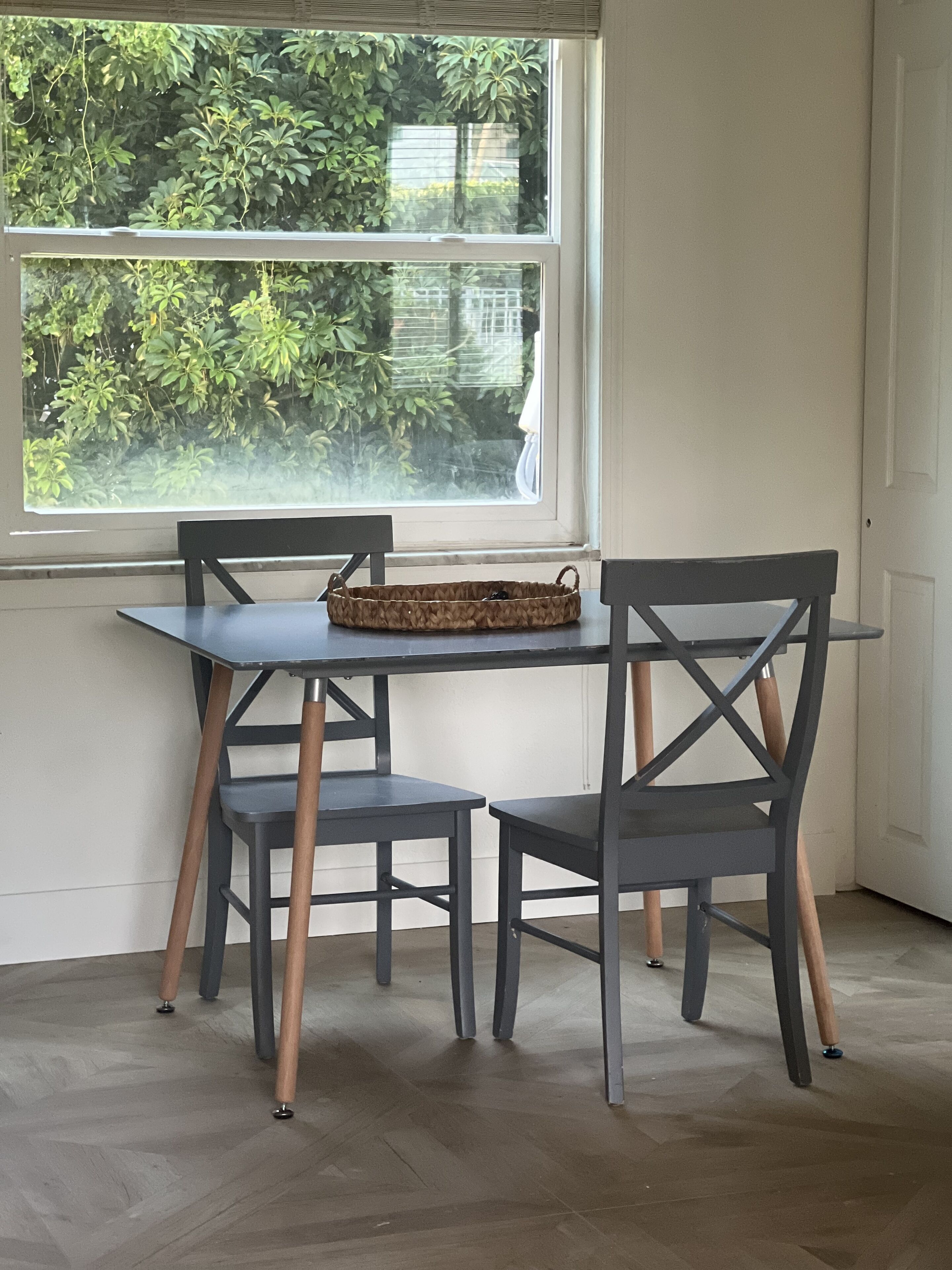 A dining area with a table and chairs, a woven basket on the table, and a view of greenery through a window.