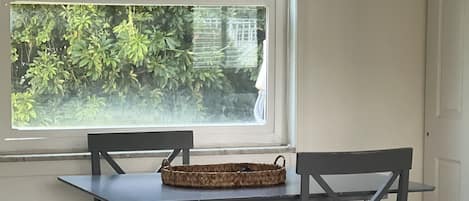 A dining area with a table and chairs, a woven basket on the table, and a view of greenery through a window.