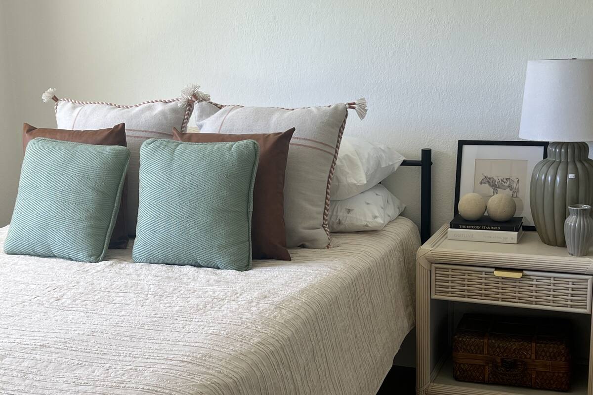 Ein ordentlich gemachtes Bett mit weißer Steudecke, zwei türkisfarbenen Kissen und mehreren braunen Kissen. Neben dem Bett steht ein kleiner Nachttisch mit einer Lampe, einem gerahmten Bild und einer Vase.