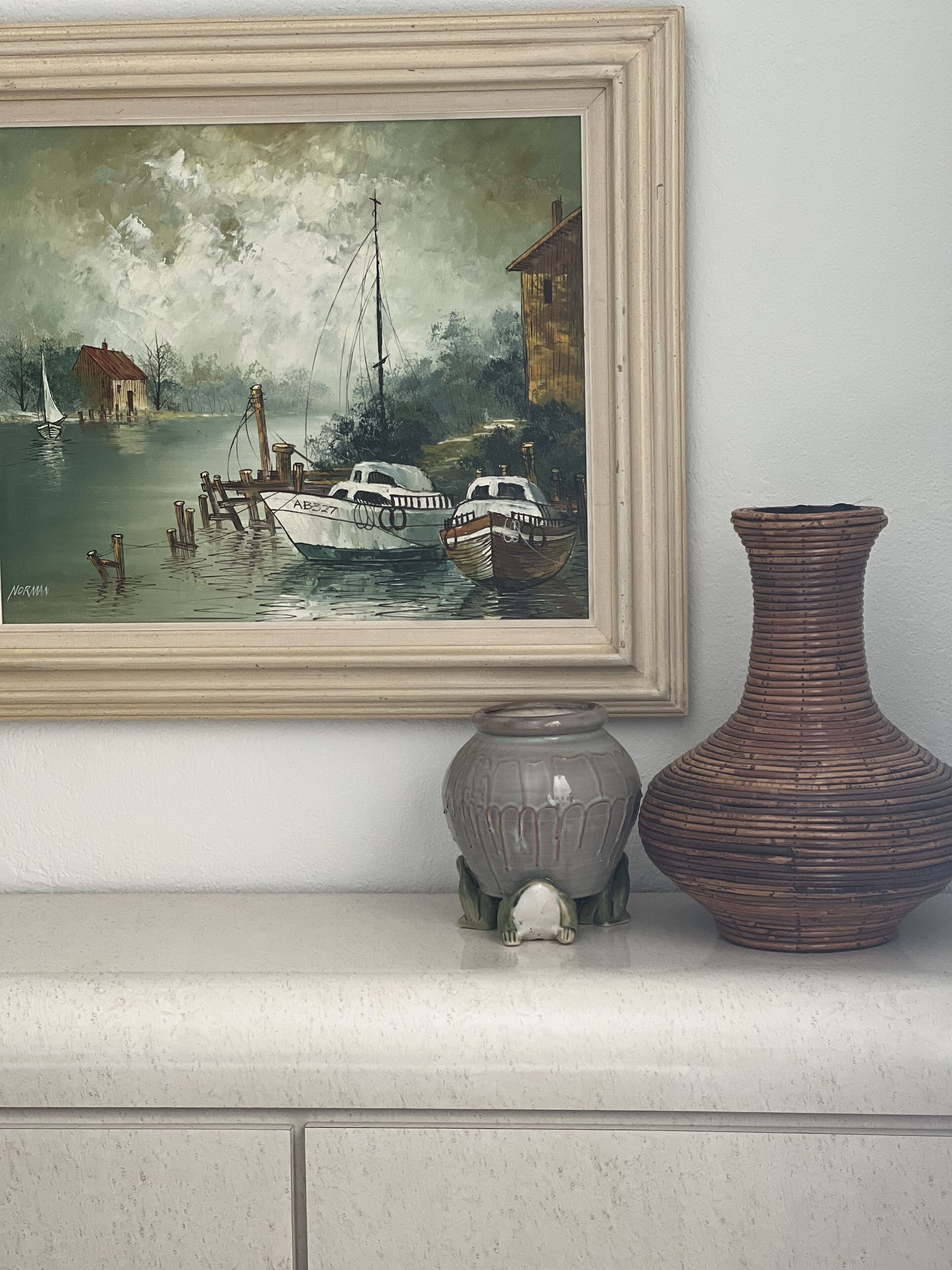 A framed painting of a harbor scene with boats, a house, and a vase on a shelf.