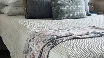 Una cama con una colcha blanca acolchada, un plaid estampado y varias almohadas de distintos colores y diseños.
