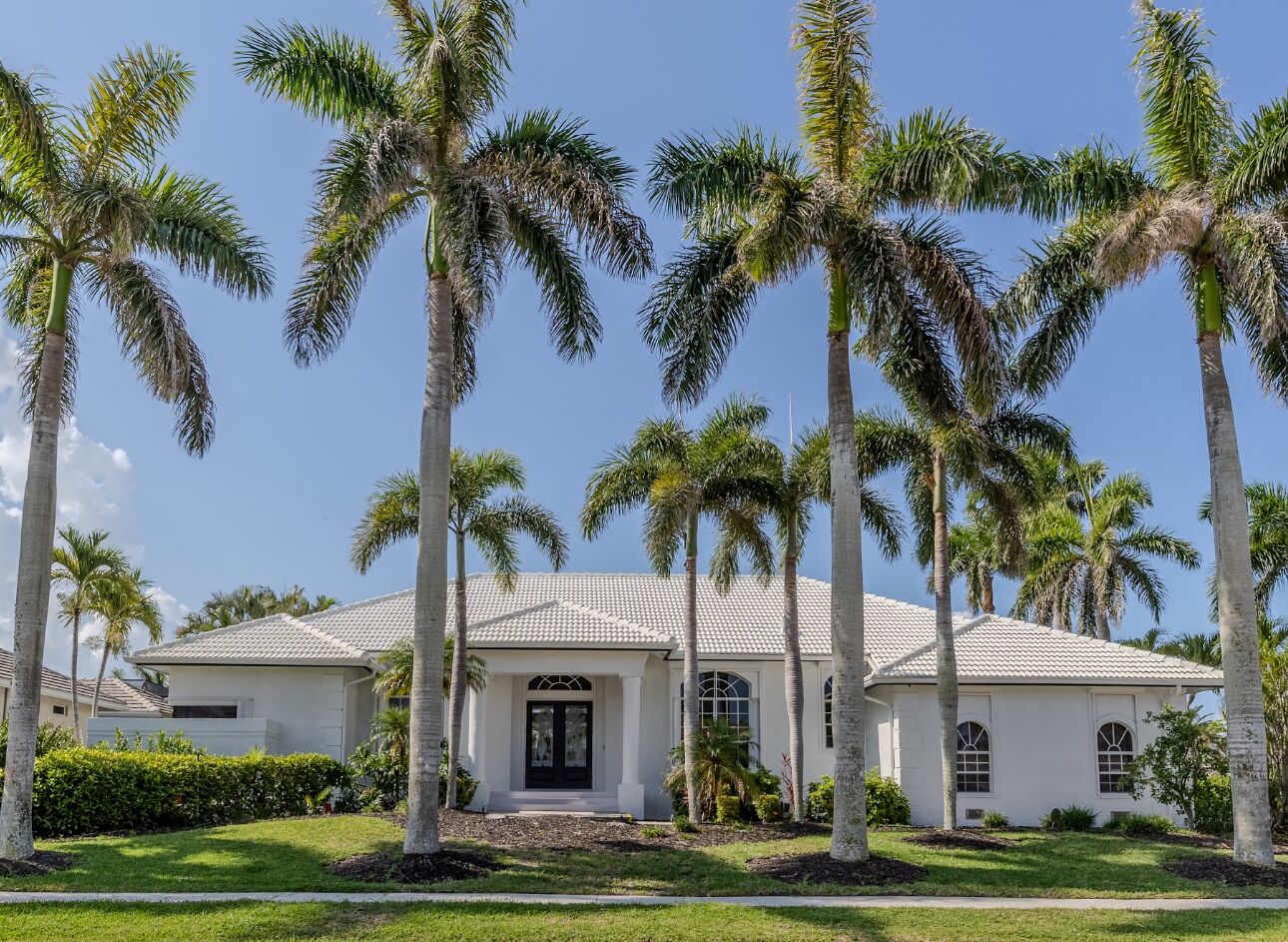 A white house with a tiled roof, surrounded by palm trees and a well-maintained lawn.