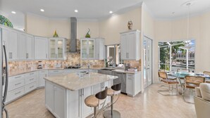 A modern kitchen with a large island, white cabinetry, and a dining area with a view of the outdoors. - Marco Island Waterfront Single-Story Home w/ Direct Gulf Access (Marco Island)
