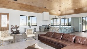A spacious living area with a leather sofa, round coffee table, and a rustic rug. A kitchen with a bar and large windows is visible in the background. - Breathtaking modern 100 acre ranch on the Colorado River. Weddings available (Smithville)