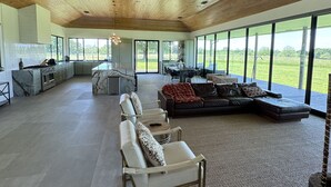 A spacious living area with a high wooden ceiling, large windows offering a view of the outdoors, a seating area with sofas and chairs, and a kitchen with modern appliances. - Breathtaking modern 100 acre ranch on the Colorado River. Weddings available (Smithville)