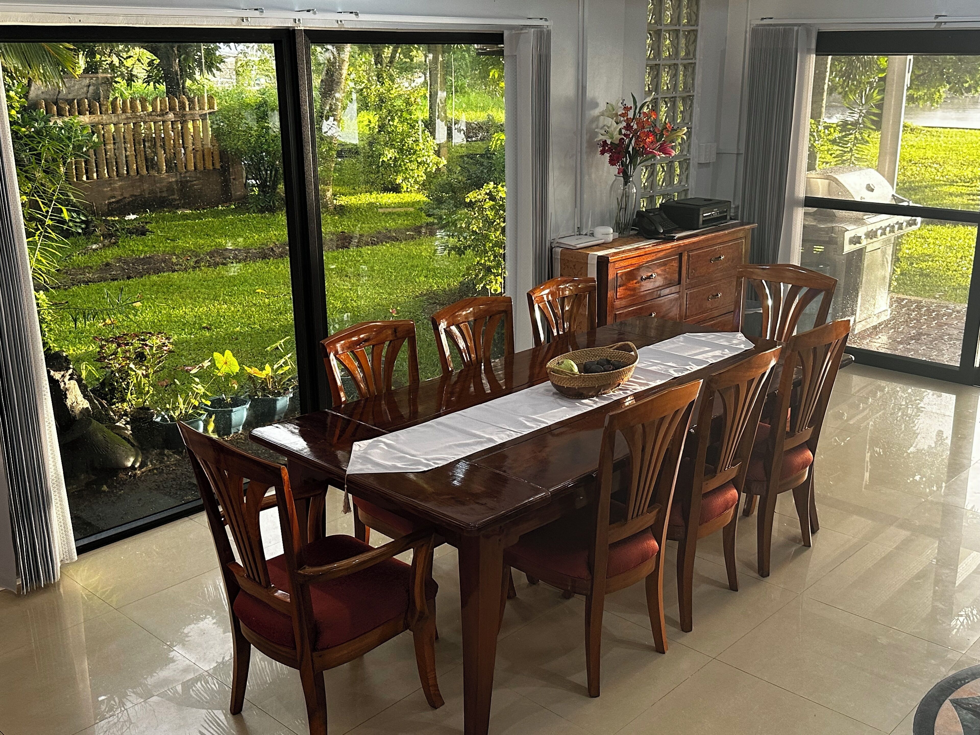 A dining area with a table set for six, surrounded by wooden chairs with red cushions. A wooden cabinet with drawers is against the wall, and a vase with flowers is on the table.