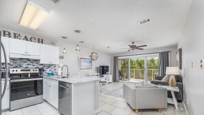 A modern kitchen with white cabinets, a built-in oven, and a central island. There is a dining area with a grey sofa and a ceiling fan. - Private Beach Access! Complementary Beach Service! (Miramar Beach)