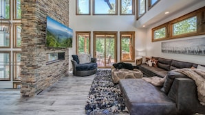 A spacious living room with a stone wall, large windows, a ceiling fan, and a fireplace.