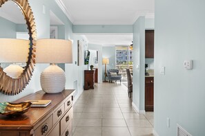 A hallway with a wooden dresser, a large mirror, and a lamp. There is a blue sofa and a dining table in the background.