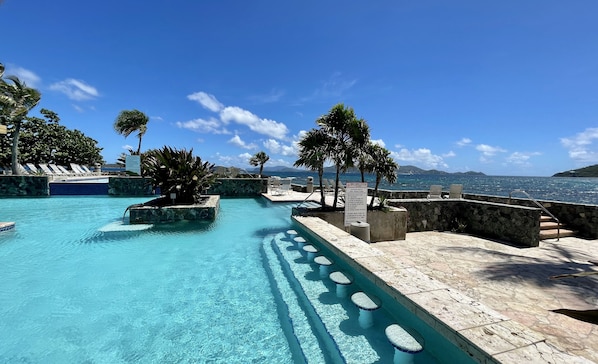 A clear blue pool with steps leading to a lounge area with chairs and a view of the sea.