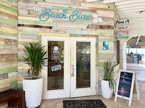The exterior of The Beach Buzz, a coffee, sundries, and ice cream shop with a rustic wooden facade, a glass door, and signage indicating the direction to the bar and food.