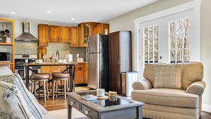 A modern kitchen with wooden cabinets, a stainless steel refrigerator, and a dining area with a bar stool. - Shamrock by the Sea Hot Tub Arcade Basketball Court & Beach Fun (Wells)