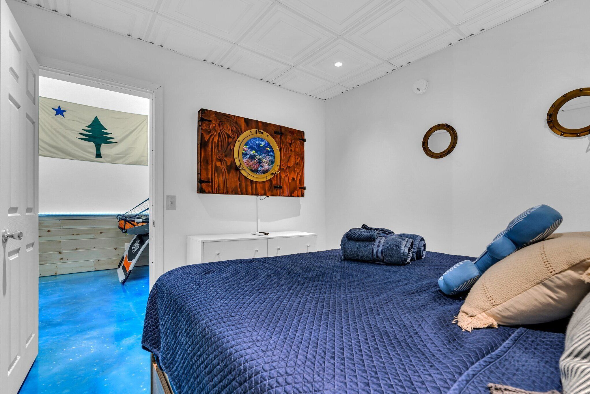 Un dormitorio con cama, un espejo circular sujeto a la pared, una bandera y una puerta que da a una habitación con piso azul.
