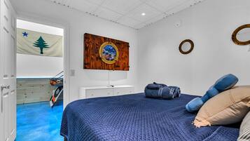 Un dormitorio con cama, un espejo circular sujeto a la pared, una bandera y una puerta que da a una habitación con piso azul.