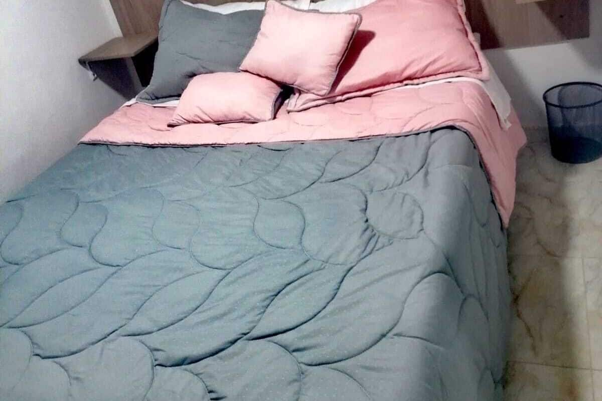 A bed with a grey quilt and pink pillows, a wooden headboard, and a small bedside table with a lamp.