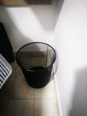 A black mesh trash bin on a tiled floor next to a white wall. - hotel 51b (Bogotá)