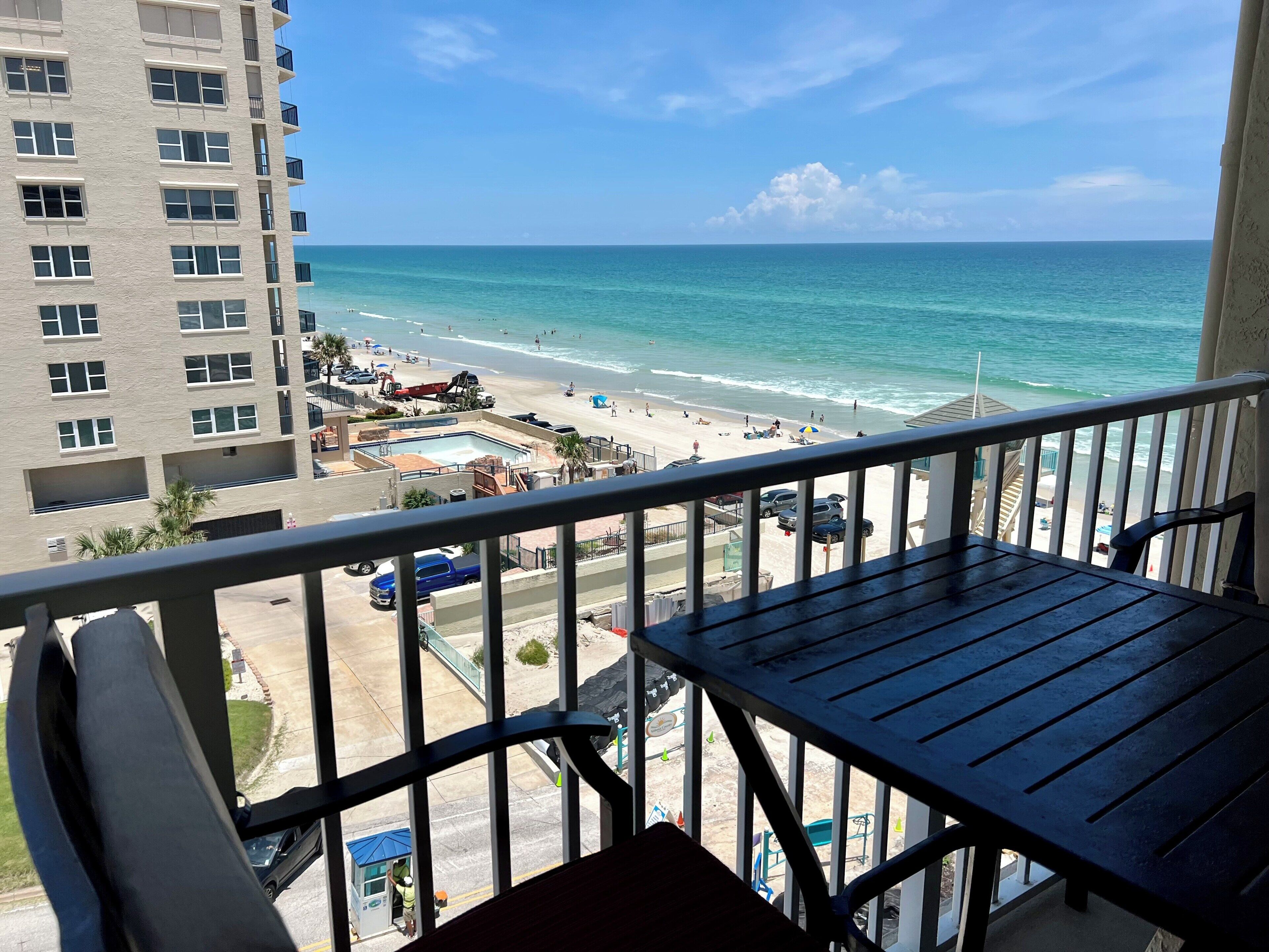 Ein Balkon mit Blick auf einen Strand und das Meer. Es gibt einen Poolbereich mit einem Rettungsschwimmturm und ein mehrstöckiges Gebäude mit Balkonen.
