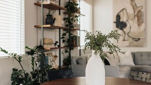 A dining area with a round wooden table, a white vase with greenery, and a grey sofa in the background. - Brand NEW4BR, Views, Family Friendly (San Diego)