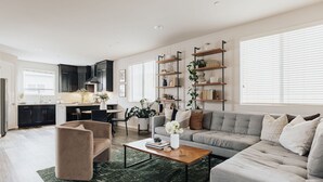 A modern living room with a grey sofa, a beige armchair, and a coffee table. There is a kitchen in the background with dark cabinets and a window. - Brand NEW4BR, Views, Family Friendly (San Diego)