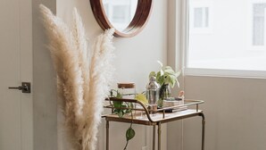 A room with a white door, a round mirror, a white vase with pampas grass, a metal cart with various items, and a window with a view of buildings. - Brand NEW4BR, Views, Family Friendly (San Diego)