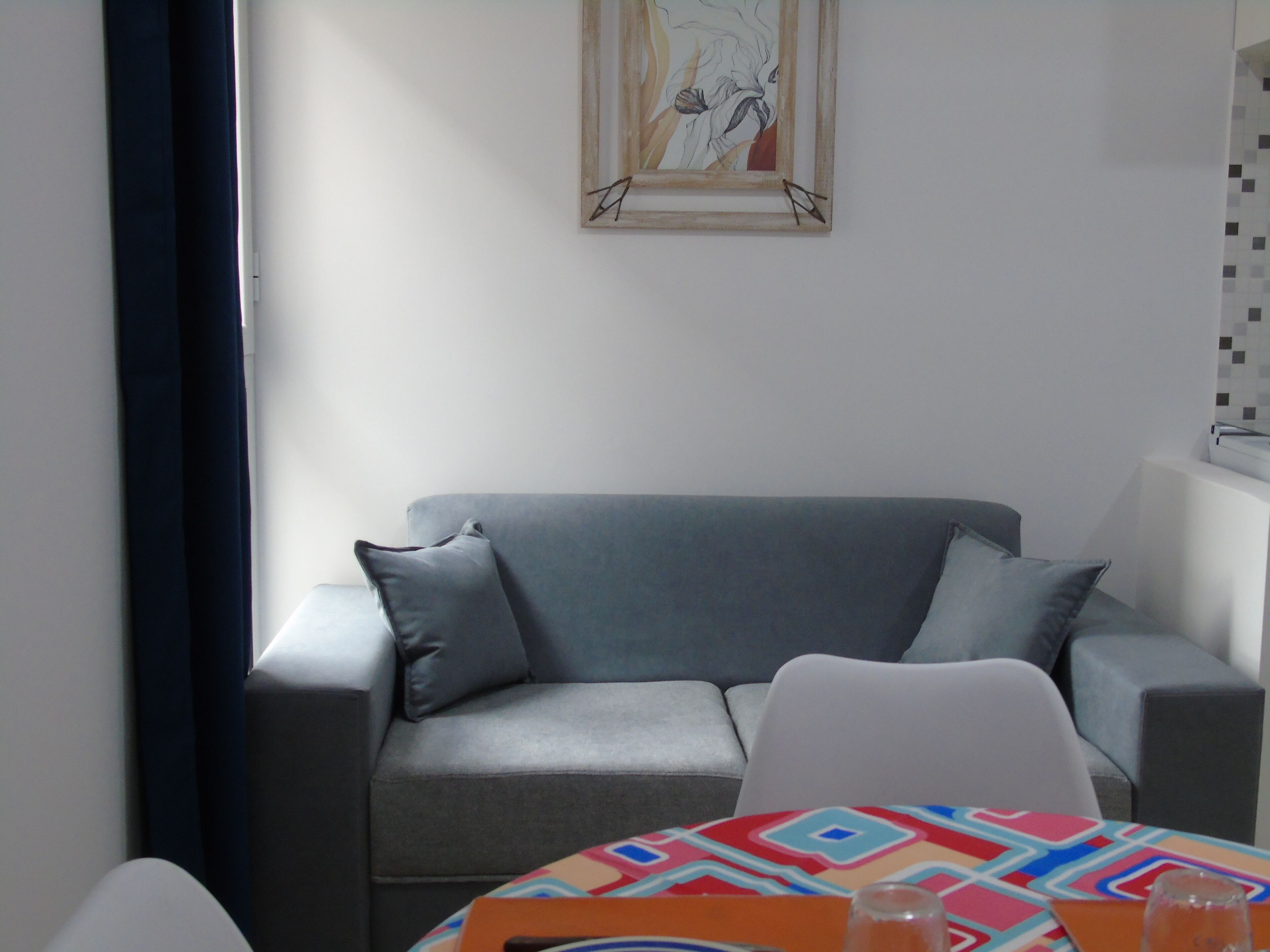 Een moderne woonkamer met een grijze bank, een witte stoel en een kleurrijke tafel. Een ingelijst kunstwerk hangt aan de muur boven de bank.