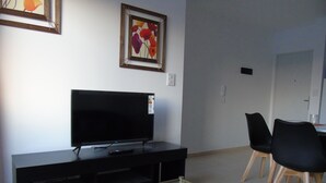 Una sala de estar con un televisor de pantalla plana sobre un mueble negro, una mesa de centro de vidrio, dos sillas negras y una obra de arte enmarcada en la pared.