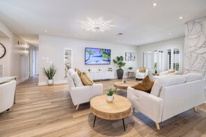 Un salon moderne avec des canapés blancs, une table basse ronde en bois et un téléviseur diffusant une scène de plage.